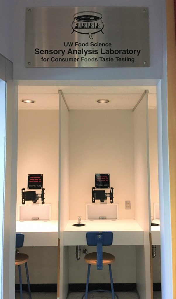 Photo shows interior of sensory analysis laboratory with two chairs at counters with a separating panel between them. Computer screens and a door from which food is served are above the counters.