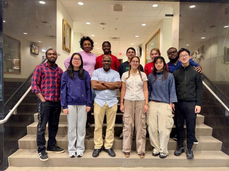 Group of people stand on the steps smiling at the camera. Man in front has arms cross, he is the PI.