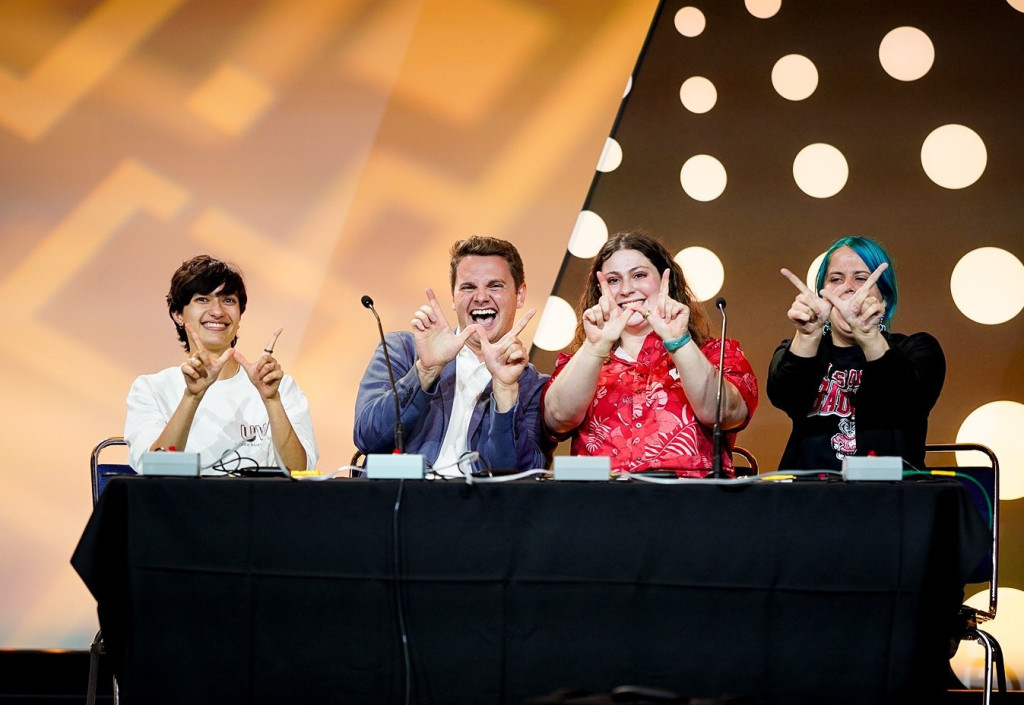 Four people sitting behind a table with a black table cloth, hold their thumb and pointer fingers in a "W" shape.