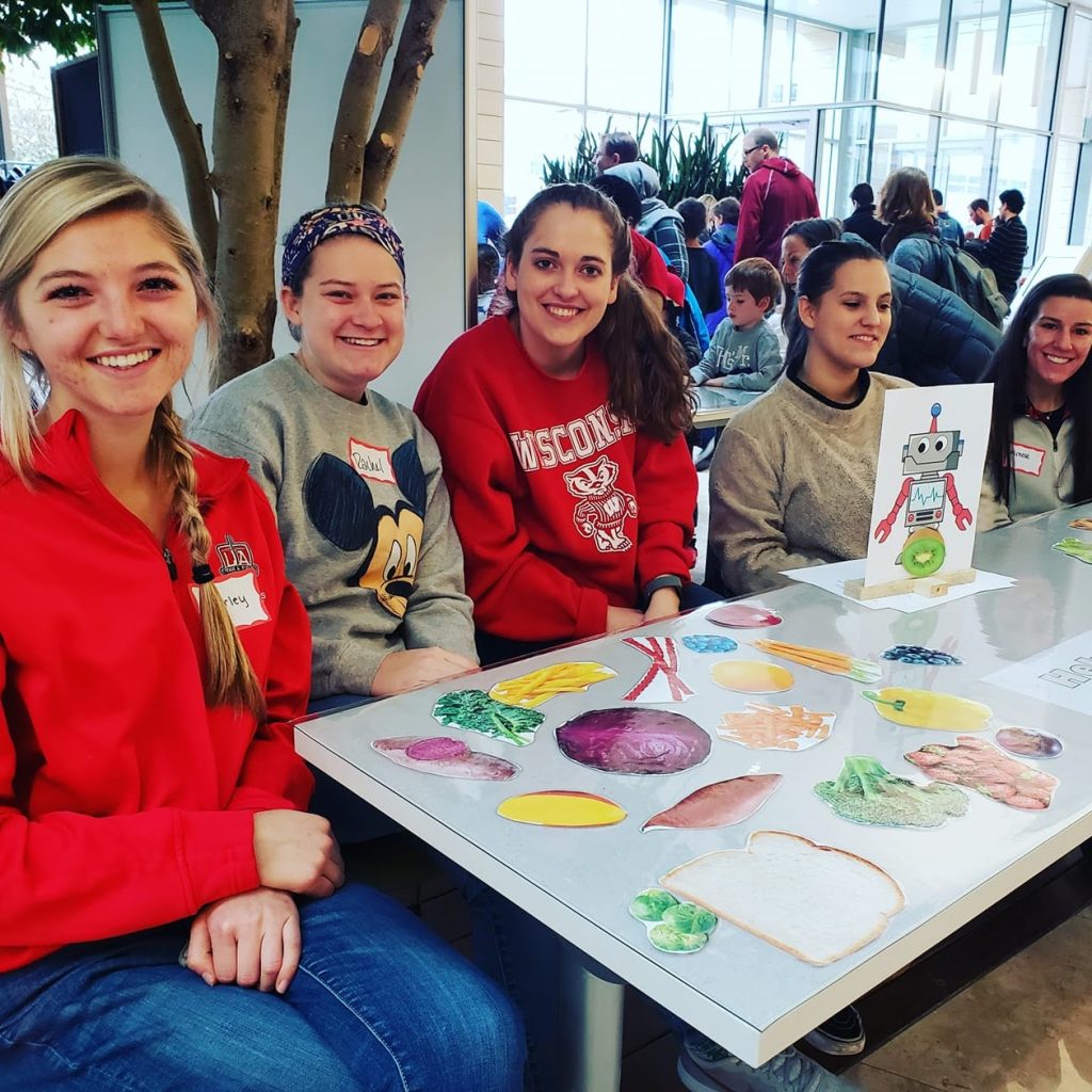 Five women sit at a table with paper food on the table.