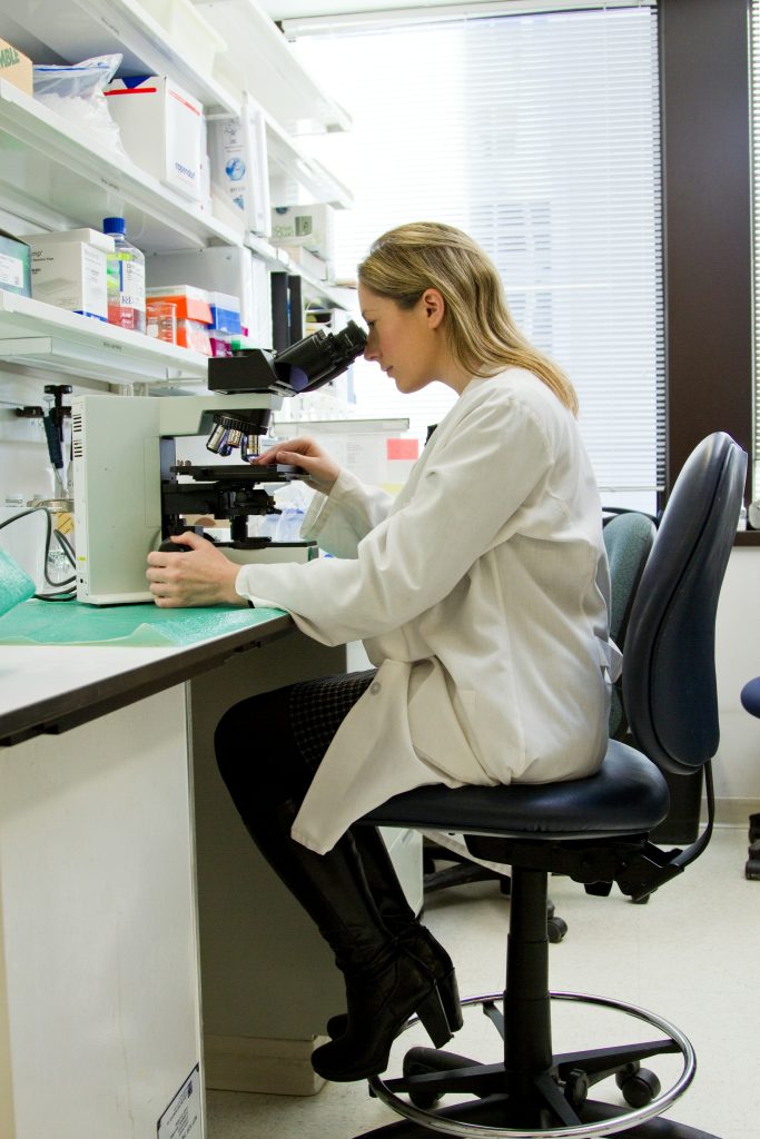woman in labcoat sitting on chair looking through microscope