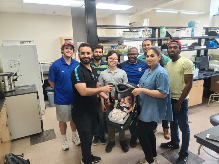 Group of people stand smiling at the camera, while lady in front holds baby in a baby carrier.