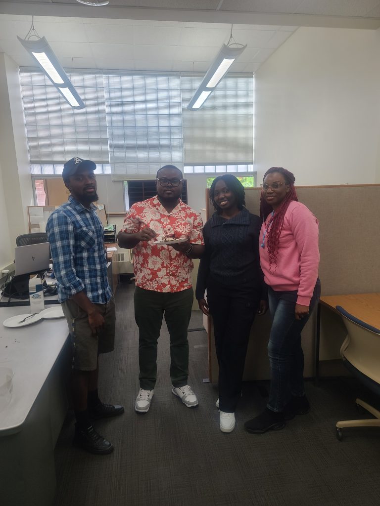 Four people stand smiling at the camera in a office setting, celebrating a birthday.