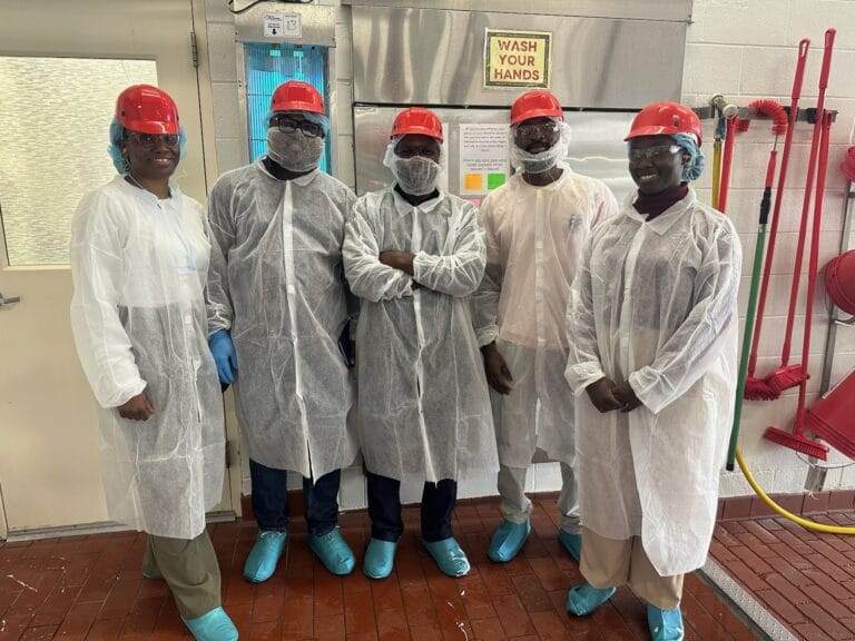 Group of five people stand side by side in a cheese factory. They are all wearing PPE.
