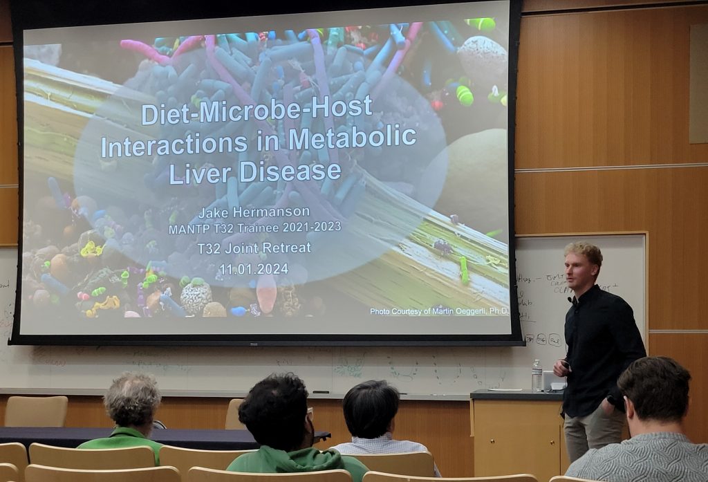 Man standing in front of a classroom, presenting his research of Diet-Microbe-Host Interactions in Metabolic Liver Disease.