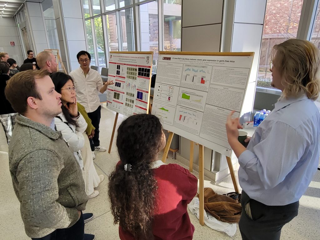 Group of people standing around a poster, listening to the speaker explain their research at the 2024 MANTP Joint Retreat.